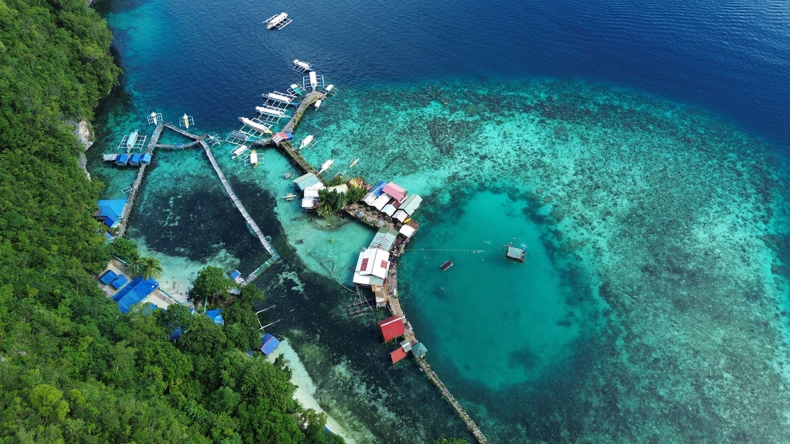 An island resort seen from above: jetties, huts on stilts and moored outrigger boats over clear reef