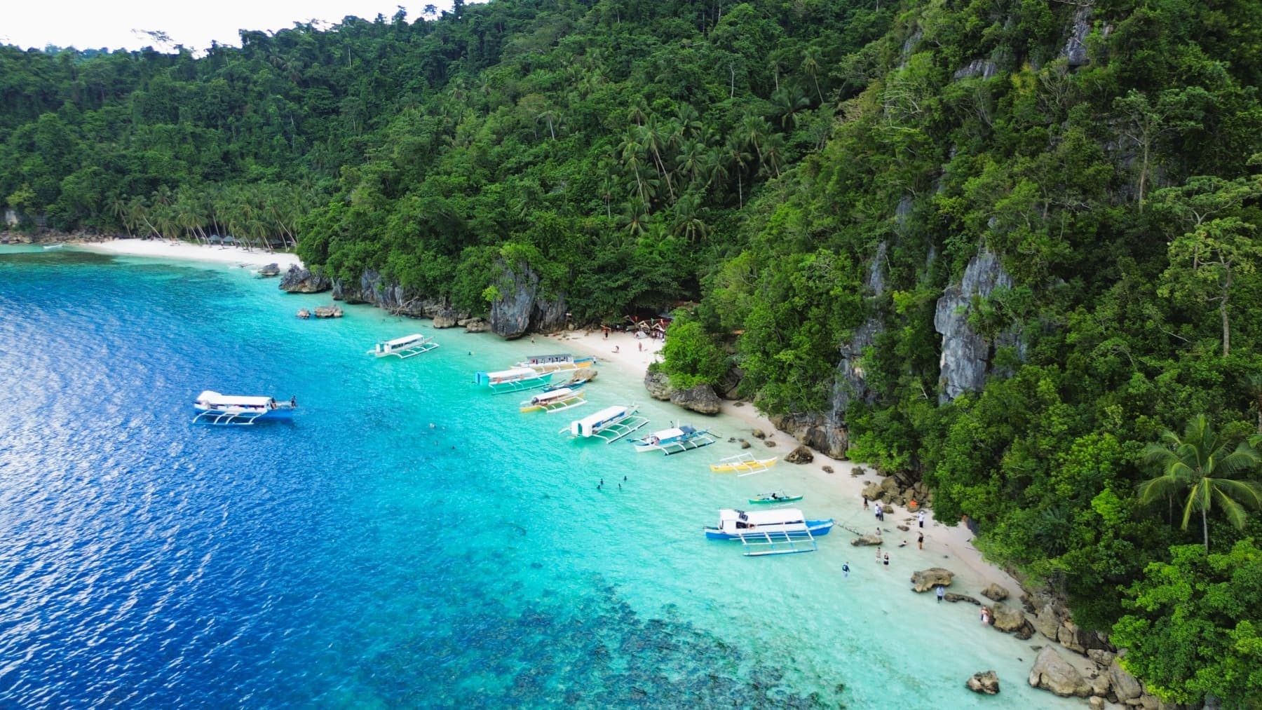 Outrigger boats drawn up on a white sand beach at Bitaog, backed by jungle and limestone cliffs