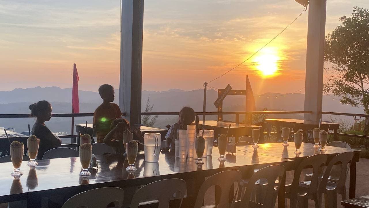 Guests seated at a long table on an open restaurant deck as the sun sets over the islands